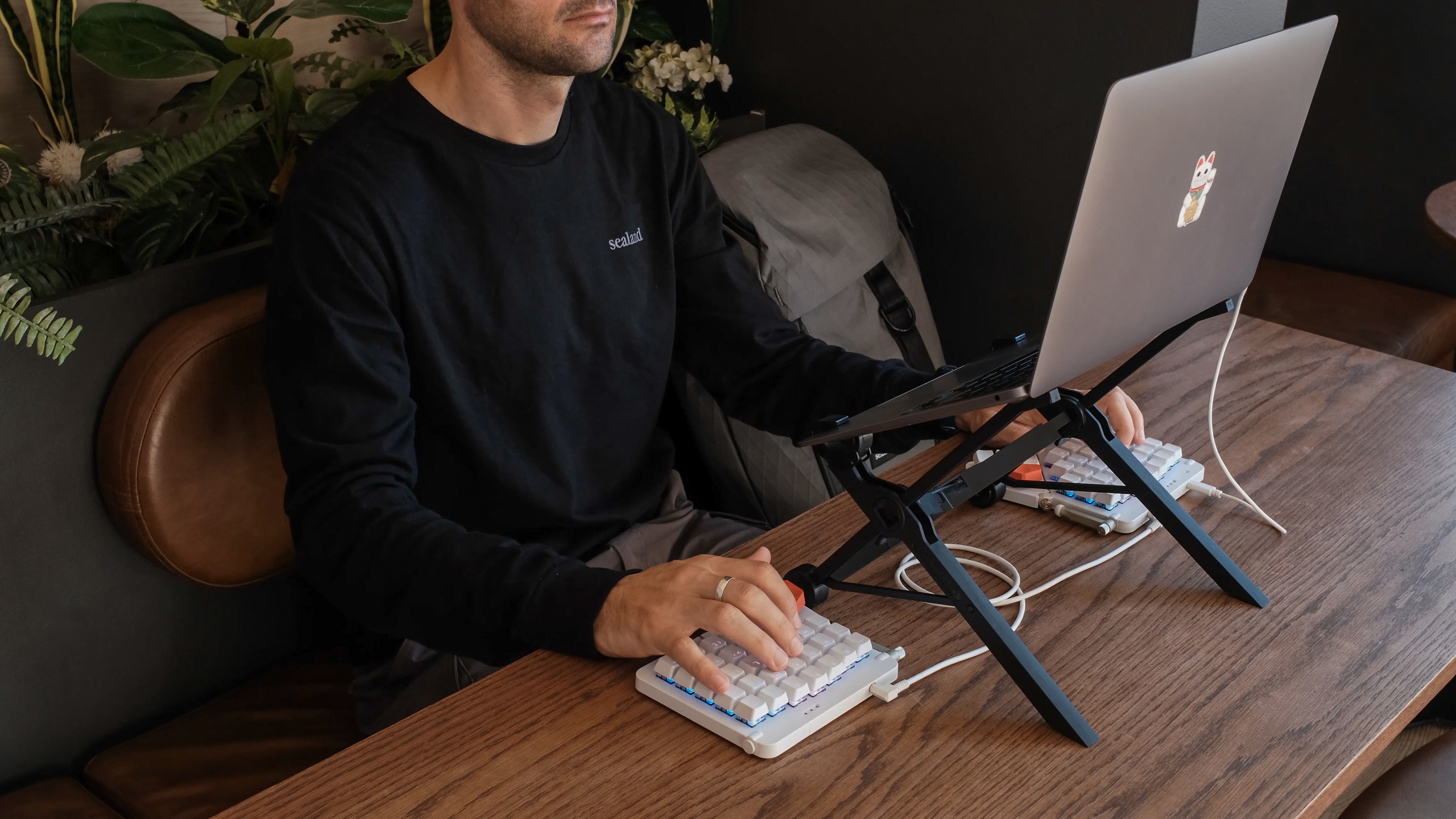 A person at a café table typing on a split keyboard, their laptop raised to eye level on the Nexstand laptop stand