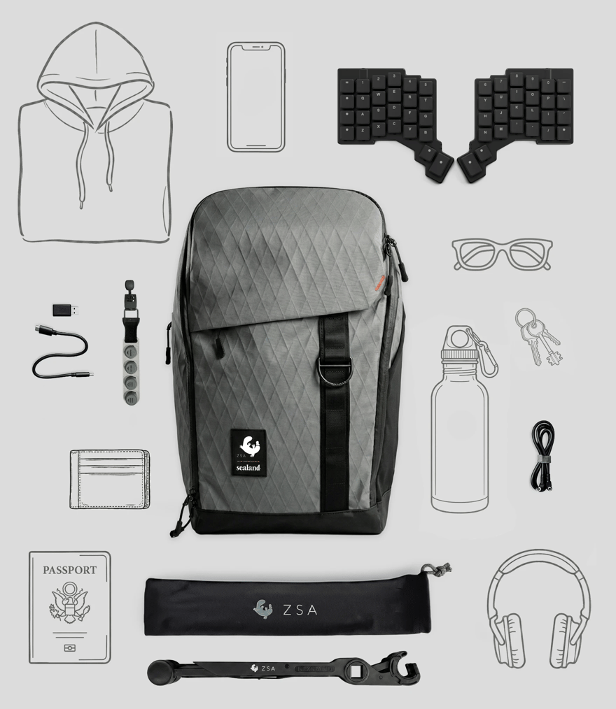 Flat-lay of the ZSA Backpack surrounded by the gear it carries: a split keyboard, laptop stand, water bottle, headphones, cables, passport, and more.
